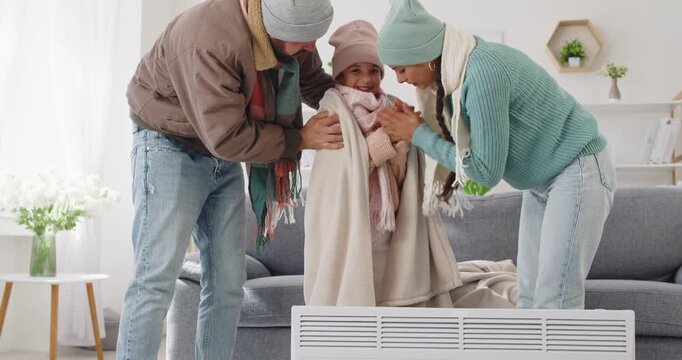 Family faces cold winter frost, seeking heating warmth. Parents help a child warm hands by a heater at home in room during a power outage noting safety and comfort. Warmth and safety at home.