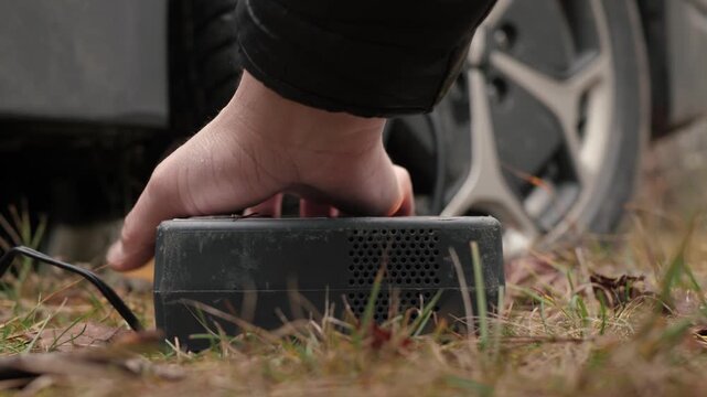 Inflating a car tire, close-up of a hand turning on a portable compressor, scene depicting turning on and servicing an air pump next to a parked car