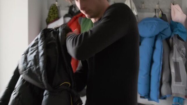 caucasian man hallway choosing winter coat, tying headband, sliding on puffer jacket, checking pockets, grabbing backpack, colorful coats on hooks, morning light, focused casual commute prep