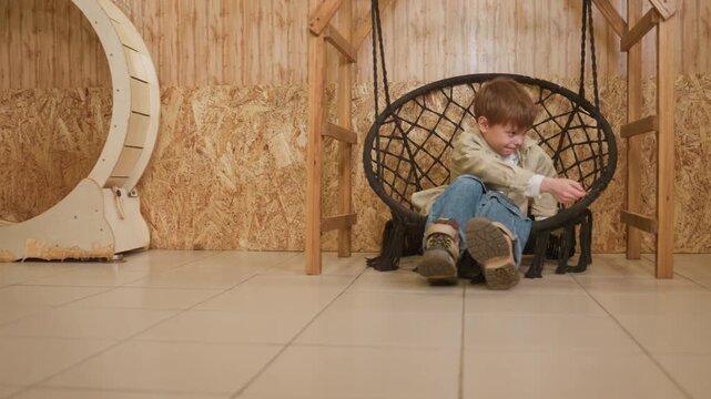 boy sitting in hanging chair reaching, indoor playroom with tiled floor and wooden wall, wicker swing chair, child stretches hand toward toy on floor, focused smile, playful determination, denim jeans