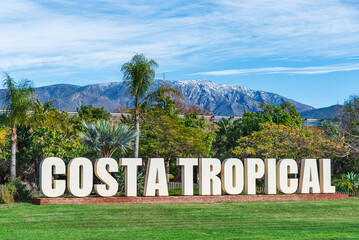 Decorative sign with the words Costa Tropical among trees and palm trees, with snowcapped mountains in the background, Motril, Granada, Spain © M. Perfectti