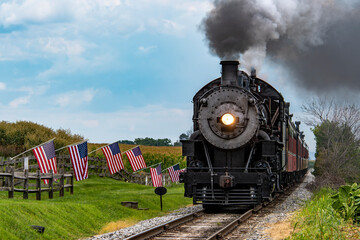 Fototapeta premium Historic Steam Locomotive Travels Along Countryside Tracks With American Flags Waving in the Breeze