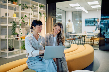 Two businesswomen discussing work using a laptop in a modern office lounge area, showing teamwork...