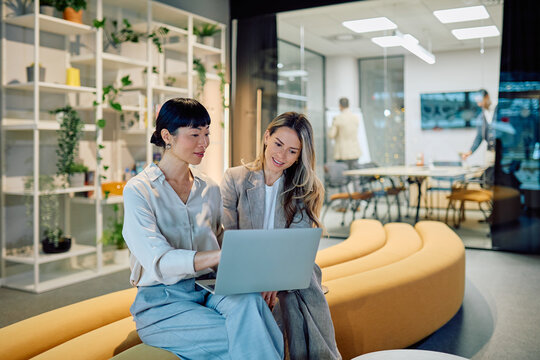Two businesswomen discussing work using a laptop in a modern office lounge area, showing teamwork and communication