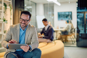 Fototapeta premium Smiling businessman wearing glasses, using a digital tablet while relaxing in a modern office break area