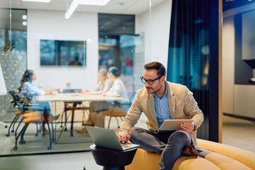 Businessman sitting on a couch, multitasking with a laptop and a tablet in a contemporary office environment
