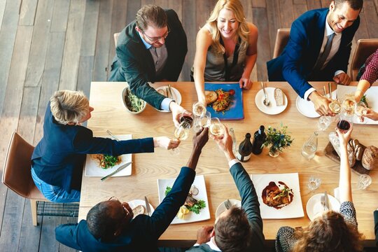 A diverse group of professionals, including men and women of various ethnicity, enjoy a celebratory dinner and toasting. The table is filled with food and drinks, highlighting a joyful gathering.