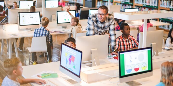 Diverse classroom with children using computers. Teacher assists students. Kids learning with technology. Engaged children in a modern educational setting. International students working on computers.