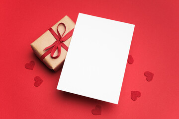 Blank Valentine's Day greeting card with copy space beside a wrapped gift adorned with a red ribbon and scattered heart-shaped decorations on a vibrant red background