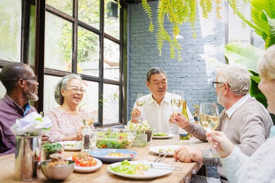 A diverse group of elderly friends, including men and women, enjoying a meal together. Elderly friends laughing, sharing wine, and dining in a cozy, sunlit room. Wine lunch fore retired men and women.