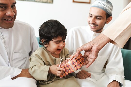 Child receiving a gift, smiling with joy. Family moment, gift-giving, happiness. Child, gift, joy. Family, happiness, gift. Smiling child, joyful moment. Muslim boy receiving a gift from family.