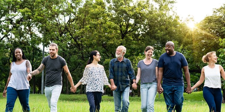 Diverse group of friends hold hands at park. Diverse group of men and women standing together hold hands at park. Diverse group of friends, men and women, standing in row hold hands on grass.