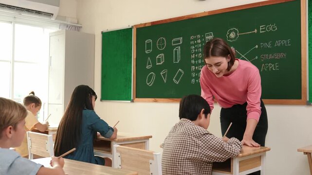 Smart teacher checking cute student homework while explaining idea at class. Attractive instructor walking around classroom while looking at notebook and talking about Math theory to child. Pedagogy.