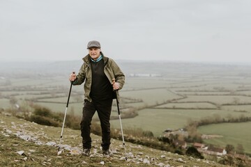 Obraz premium Elderly man hiking with poles on grass hill. Outdoor activity in nature. Senior enjoying hiking in nature landscape. Scenic landscape. Senior man hiking, outdoor activity in nature grass hill