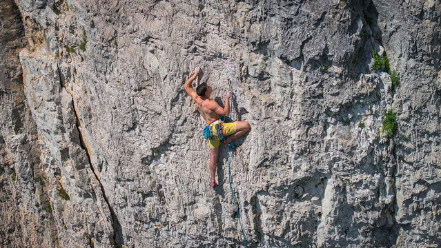 Young man lead climbing a grey limestone face in the Karst region. He is shirtless and focused, reaching to clip his rope into a quickdraw as he ascends the challenging rock face in famous Osp area.