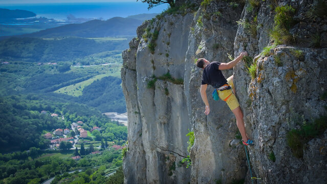 Rock climber climbs a rugged limestone cliff with a stunning view of the green karst landscape and blue Adriatic Sea on a sunny day. Freedom and challenge of outdoor climbing in famous Osp region.