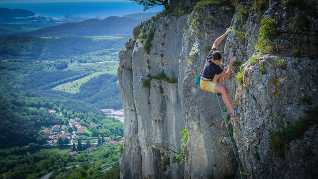 Agile rock climber searches for a foothold while lead climbing a scenic cliff in Slovenia. Below him, a picturesque village sits in green valley and deep blue Adriatic Sea is visible in the distance.