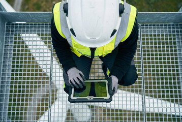 High angle view of female engineer working on digital tablet standing on metal grid platform on top of wind turbine, Industrial safety concept photography
