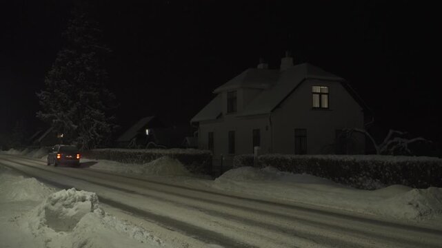 A dark minivan drives away on a cleared but snowy street at night. Large snowbanks line the road. A house with a lit window and a snow-laden tree are visible. Snow is falling in the cold dark air. 