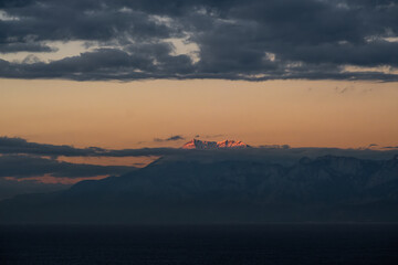 Scenic landscape of a snowy mountain peak glowing with orange sunset light, rising above layers of dark blue clouds and mist.