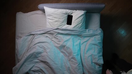 Smartphone resting on an empty bed pillow, casting a blue light glow on the duvet and wooden floor, representing sleep disruption and technology overuse