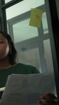 Vertical video: Flipping papers, woman in green top leading at office, writing on sticky-note glass