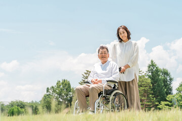 A senior man in a wheelchair in a park and a woman assisting him (caregiving, parent-child, nursing...