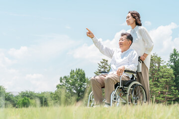 A senior man in a wheelchair in a park and a woman assisting him (caregiving, parent-child, nursing...