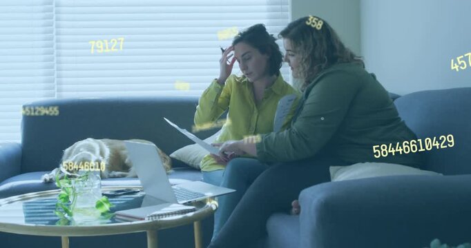 Two women arranging papers, checking laptop and comparing financial figures with numeric overlay