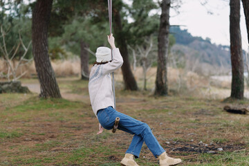 Person wearing hat and sweater swings on a rope swing in a park, surrounded by trees and open field, capturing a calm moment of relaxed outdoor recreation.