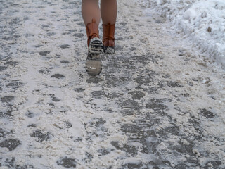 Person walking on an icy, snow-covered sidewalk during winter. Close-up of boots on slippery pavement illustrating winter hazards, pedestrian safety risks, and cold urban weather conditions