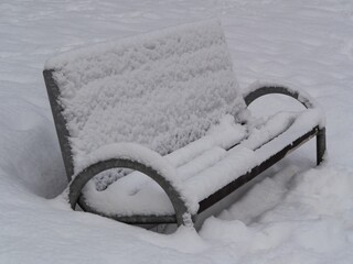 Snow-covered park bench buried in fresh snow during winter. Minimal, quiet scene symbolizing solitude, cold weather, and seasonal stillness in an urban outdoor environment