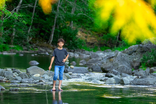 Teenager boy is standing on a rocks in the mountain rocky river with clear green waters in summer forest. Scene under branches with green lush foliage. Sooke potholes park, Vancouver island, Canada.