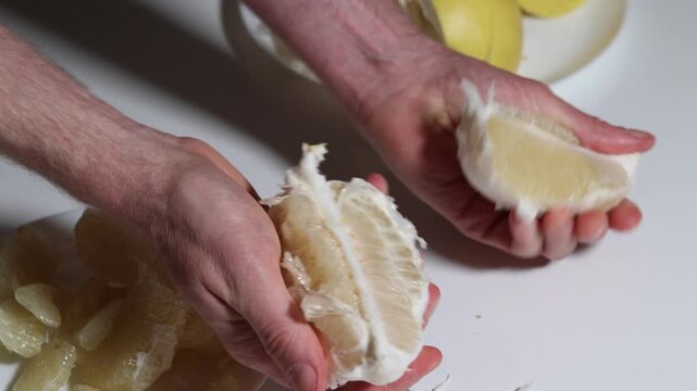 Close-up of hands gently peeling a pomelo, carefully extracting each piece against a white surface, complemented by the already-peeled pieces and leftover rinds scattered around.