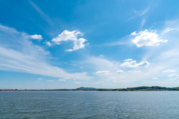 Wide landscape of a calm blue lake under a bright sunny sky with white wispy clouds. Scenic view of a reservoir surrounded by distant green hills and trees. Peaceful natural outdoor background.