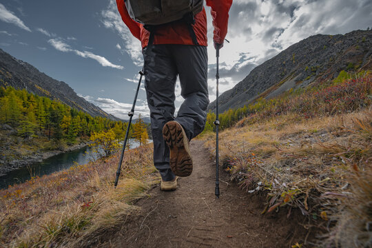 A hiker in bright gear with poles and backpack walks a forested mountain road overlooking an autumn lake. Low rear-angle shot captures boot sole and movement under a cloudy sky.