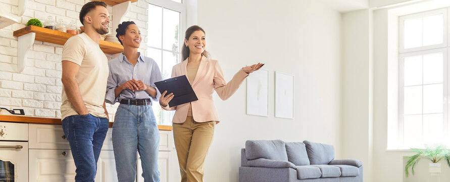 Woman realtor shows a bright apartment interior to a smiling couple as they tour rooms and discuss layout, features and the sale and ownership. Real estate agent showing new house to clients. 
