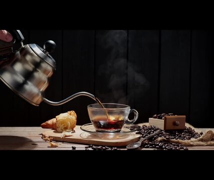 The slow-motion video shows black coffee being poured into a cup. Roasted coffee beans and bread are arranged on a table, against a black and dark background.