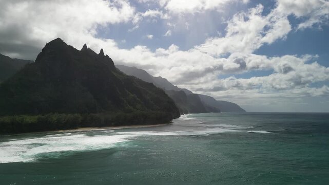 Epic aerial drone panorama of rugged Kauai coastline with dramatic emerald mountains, rolling Pacific waves, and sunlit clouds along the wild North Shore near Haena State Park
