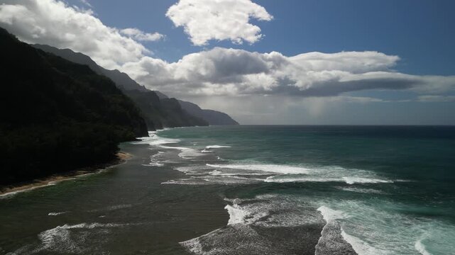 Epic aerial drone view of Kauai North Shore coastline with dramatic emerald cliffs, rolling Pacific Ocean waves, turquoise reef water, and towering clouds near Haena State Park
