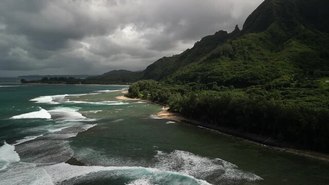 Dramatic Aerial View of Haena State Park Coastline with Lush Tropical Mountains, Dense Green Forest, Sandy Beach and Rolling Pacific Ocean Waves Under Stormy Clouds, Kauai Hawaii
