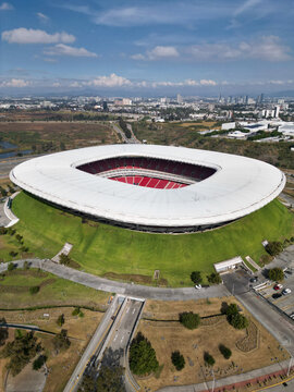 Zapopan, Mexico - December 7 2025: Captivating aerial view of modern Akron Stadium in Guadalajara. Distinctive white roof, red seats, green slopes, and urban backdrop under blue sky