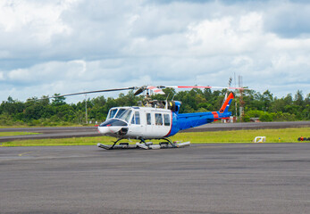 Stationary helicopter on the runway at Tanjung Pandan Airport, Belitung. An aircraft parked on the apron surrounded by green trees and a cloudy sky.