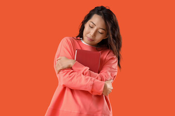 Young Asian woman with book hugging himself on orange background
