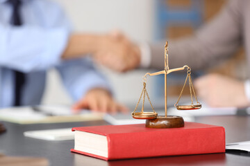 Justice scales with book on table against men shaking hands at court mediator's office, closeup