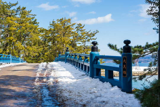 The distinctive blue railing of the Daitenkyo Bridge along the snow-dusted path of the Amanohashidate sandbar on a clear winter day. Miyazu, Kyoto, Japan. Japanese translation: Daitenkyo Bridge.