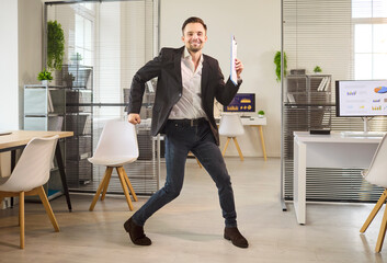 Fototapeta premium Businessman office success happy dance celebration victory. He celebrates growth with reports in a workspace, symbolizing profit, motivation and achievement. Clear concept of victory and joy.