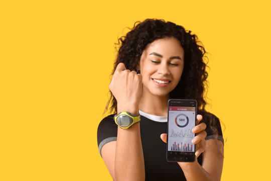 Sporty young African-American female coach holding mobile phone with calories counting app and fitness bracelet on yellow background