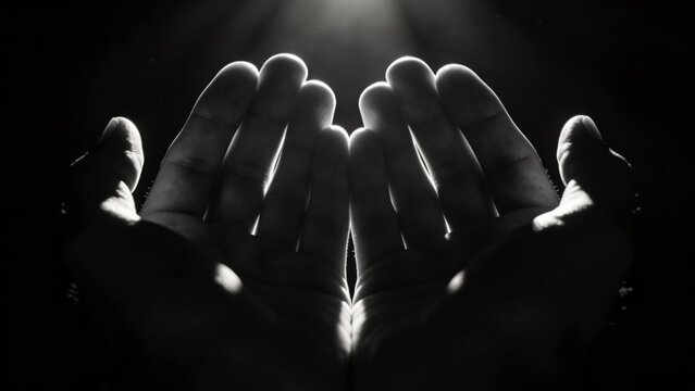 Two cupped human hands are held up receiving dramatic light rays in a dark studio setting signifying concepts of prayer, hope, and spirituality