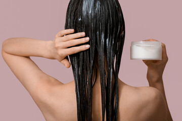 Naklejka premium Beautiful young woman with jar of cosmetic product applying hair mask on pink background, back view